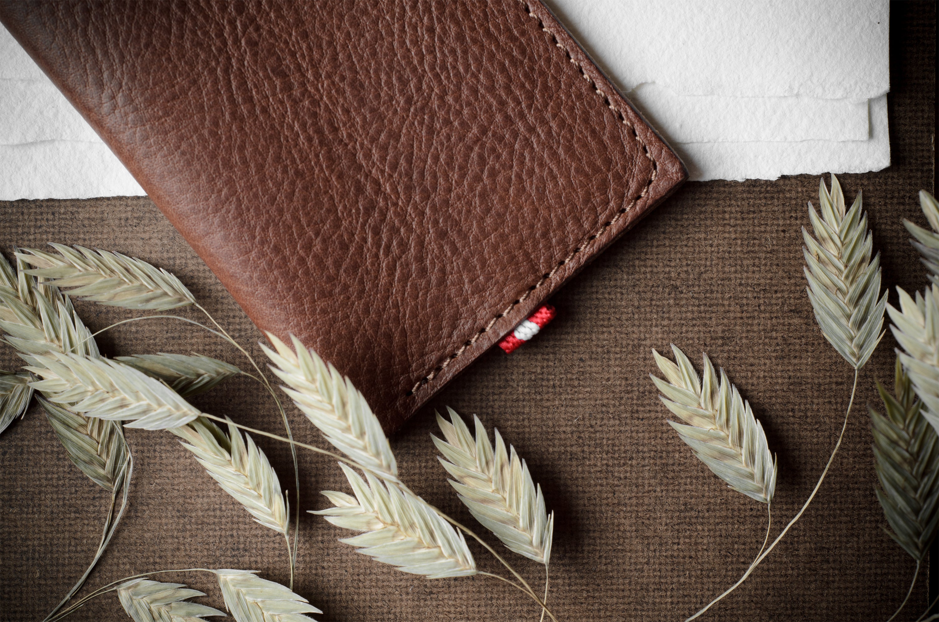 A brown textured leather card wallet, shown closed on a brown burlap-like surface with dried oat stalks and a white paper corner visible.