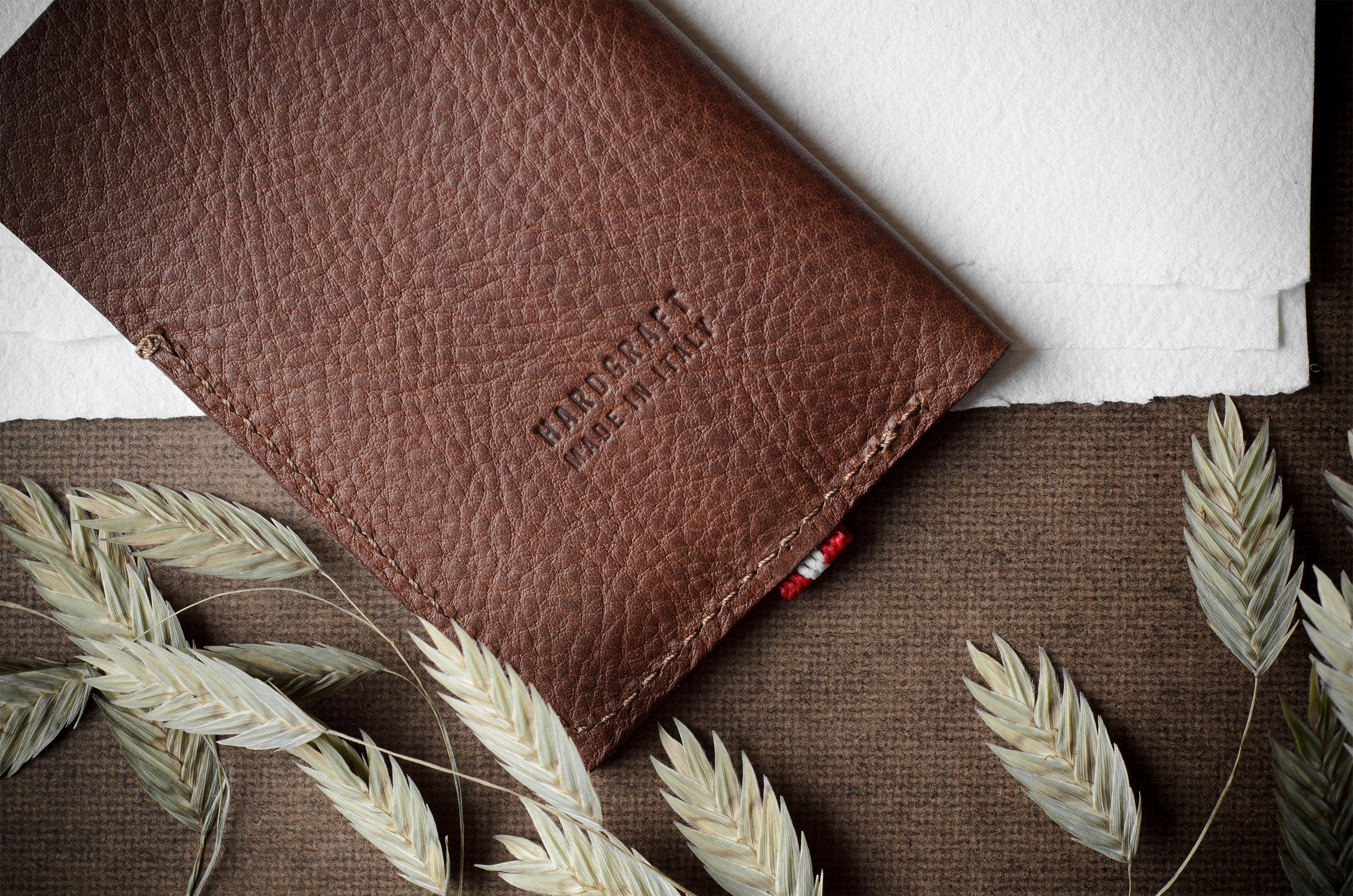 A brown textured leather card case or wallet with 'HARDGRAFT MADE IN ITALY' embossed on the front, styled flat-lay with dried oat stalks on a brown burlap surface and white paper in the background.