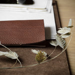 A close-up of a brown leather card case resting on a brown fabric surface, with dried white wheat-like leaves draped across it on a wooden tabletop.
