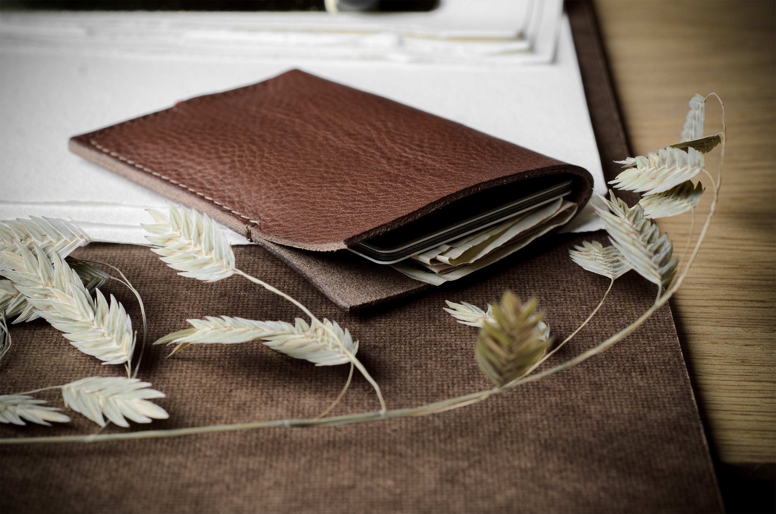 A brown leather card holder wallet with card slots, shown open with cards partially visible, styled on a brown fabric surface with dried oat grass sprigs.