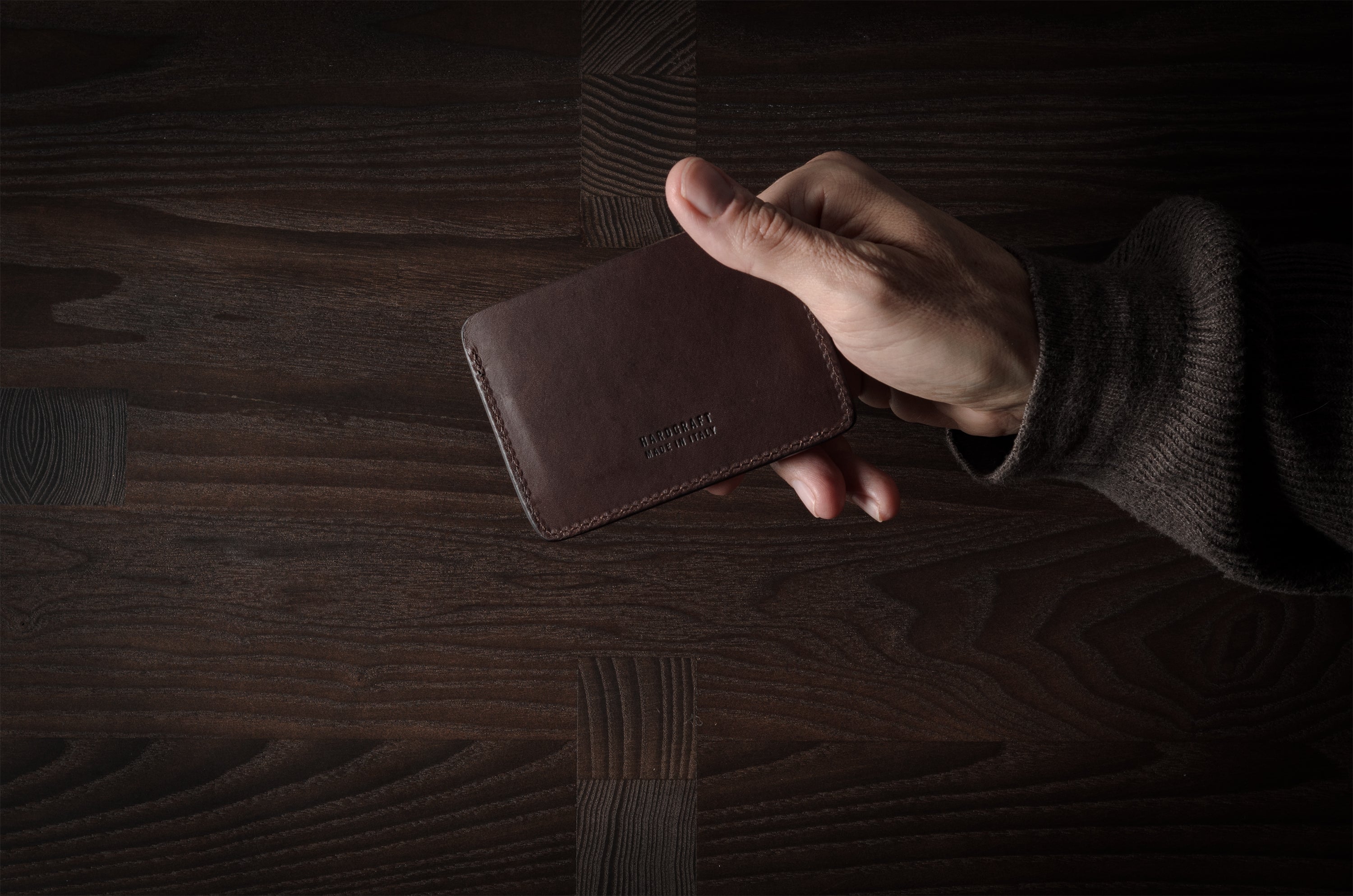 Brown leather card case held by hand on a dark wood background.