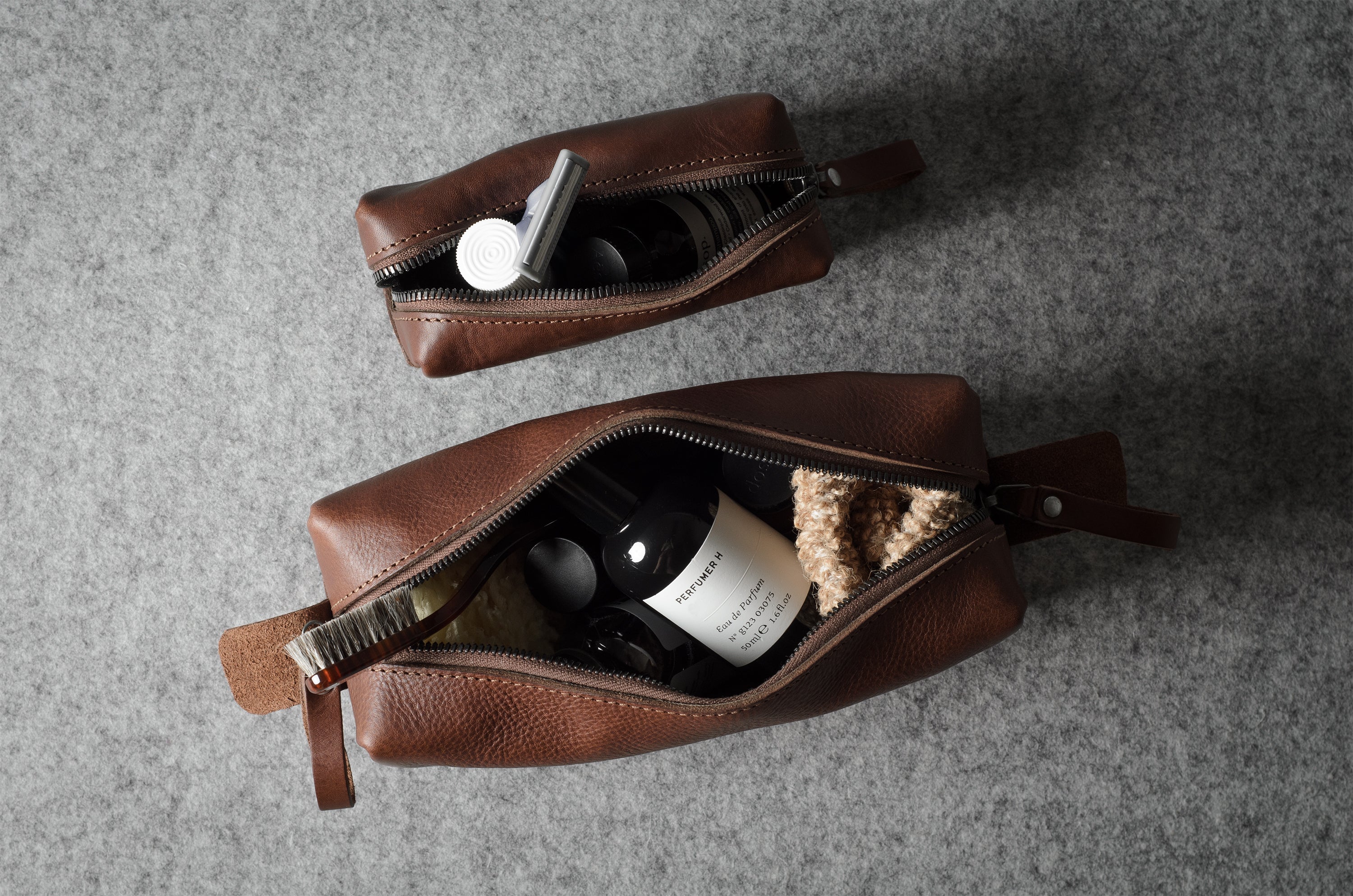 Two brown leather zippered pouches arranged on a gray textured surface, each open to show toiletry items inside.