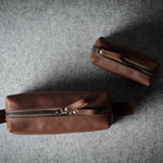Two brown leather zippered dopp-kit pouches with leather pull tabs, displayed on a textured gray felt background.