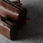 Two brown leather zippered pouches, one small and one larger, arranged diagonally on a textured gray felt surface.