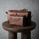 Two brown leather pouches of varying sizes stacked on a round wooden stool against a textured gray wall.