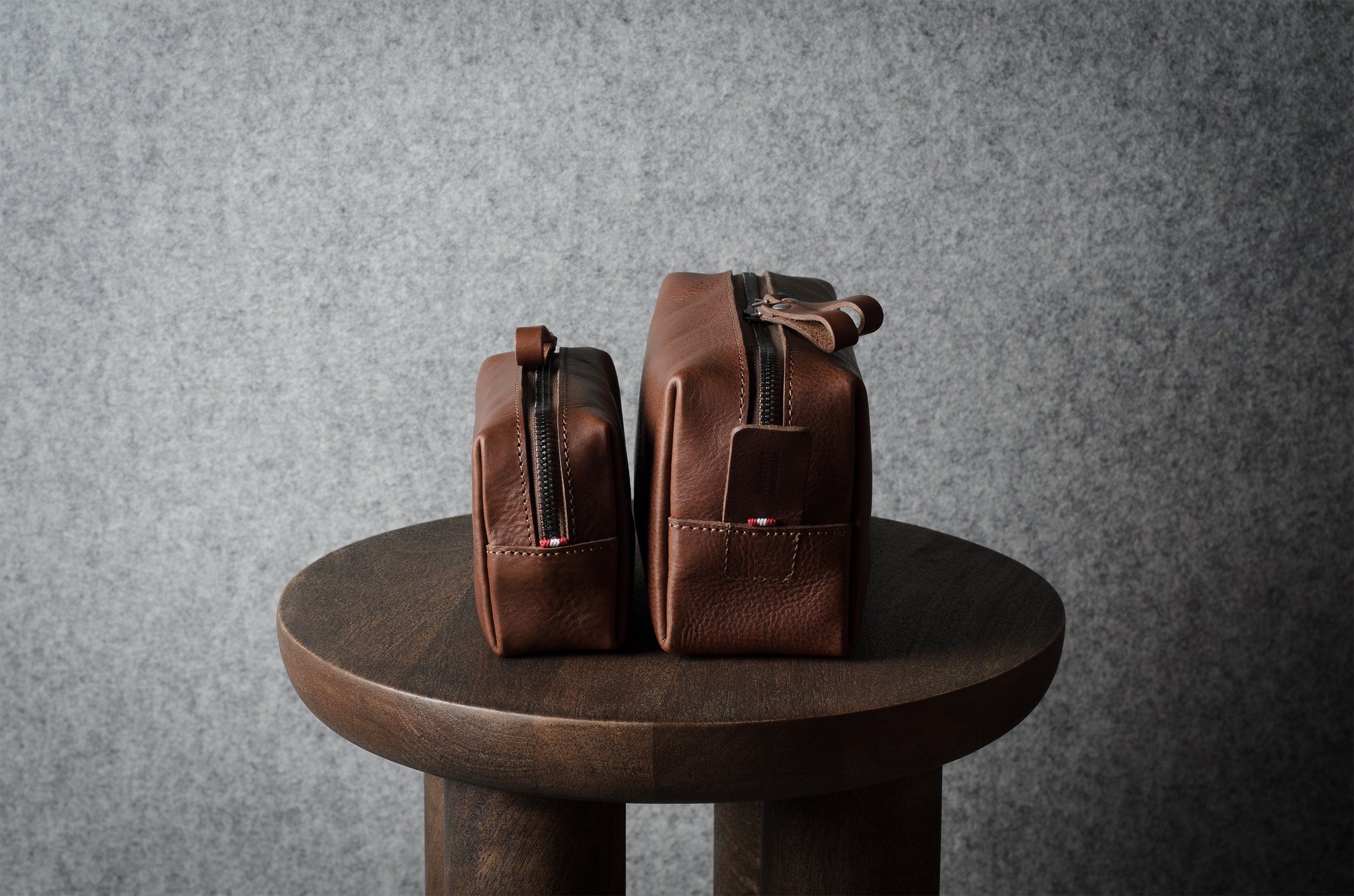 Two brown leather zippered pouches or small bags displayed side by side on a round wooden stool against a textured gray wall.