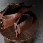 Two brown leather zip pouches with leather pull tabs, displayed on a round wooden stool against a dark gray textured background.