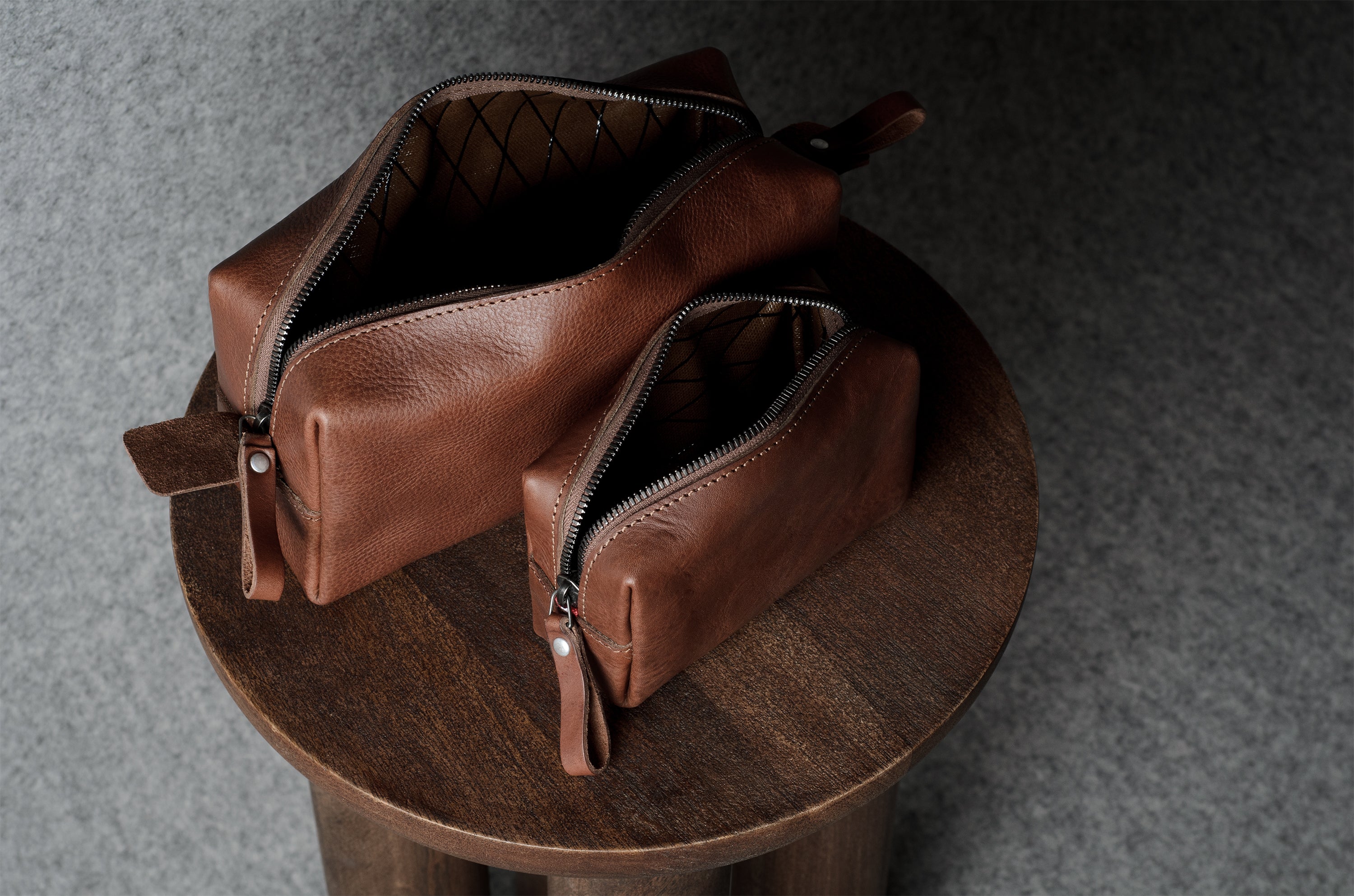Two brown leather zip pouches with leather pull tabs, displayed on a round wooden stool against a dark gray textured background.