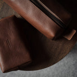 Two brown leather zip pouches, one smaller flat pouch and one larger cylindrical pouch, displayed on a round wooden surface against a gray background.