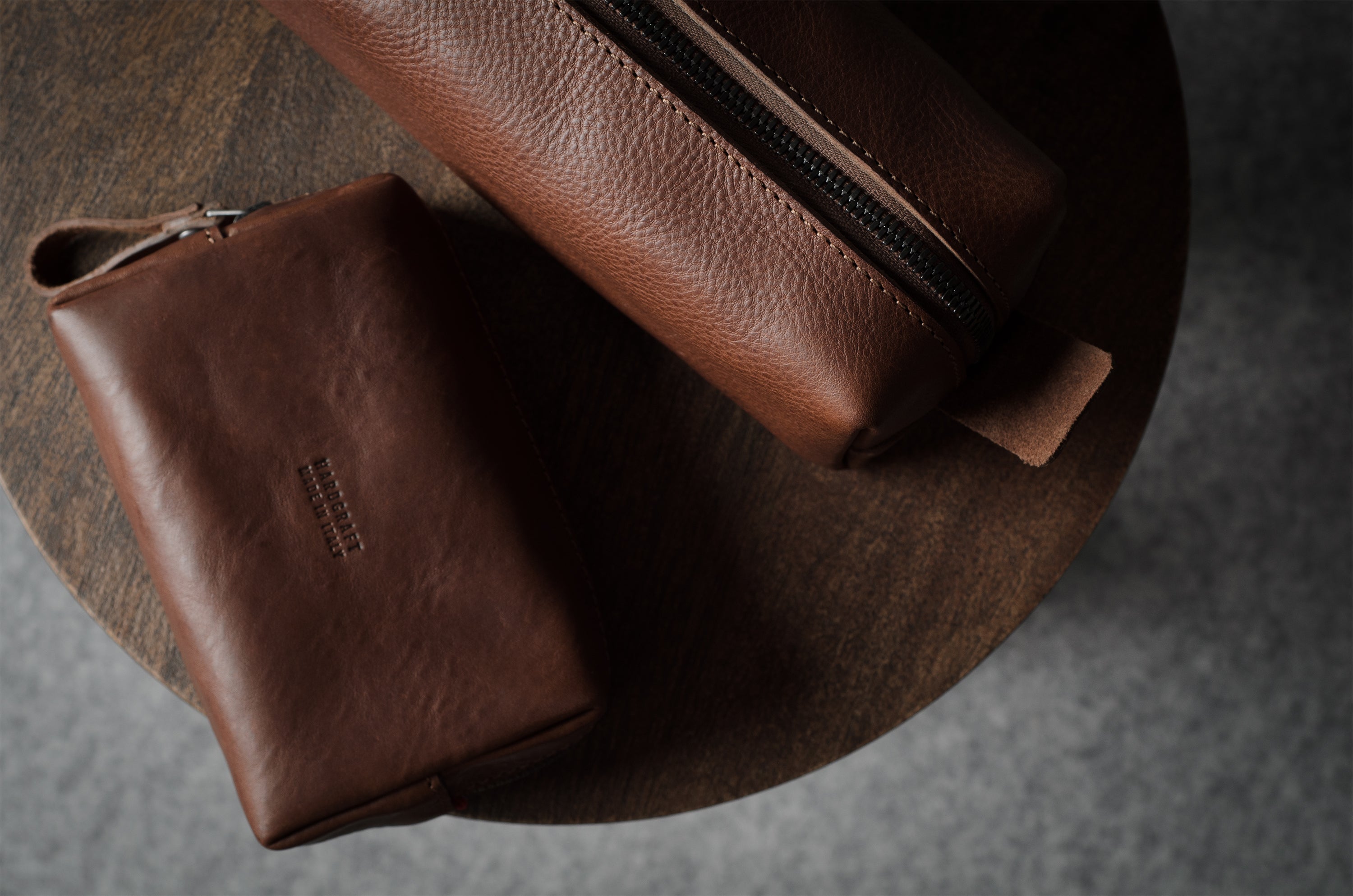 Two brown leather zip pouches, one smaller flat pouch and one larger cylindrical pouch, displayed on a round wooden surface against a gray background.
