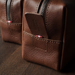 A close-up of a brown pebbled leather zippered pouch with a front pocket and a small red-and-white striped accent near the zipper, photographed on a dark wooden surface.