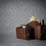 Two brown leather zippered pouches or small bags with loop handles, shown on a textured gray surface against a gray backdrop. One pouch is open, revealing a beige toothbrush and a small white item inside.
