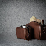 Two brown leather zippered pouches or small bags with loop handles, shown on a textured gray surface against a gray backdrop. One pouch is open, revealing a beige toothbrush and a small white item inside.