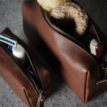 Two brown leather zippered toiletry bags on a gray textured surface, with one bag open to show a shaving razor and the other open to show a bath sponge.