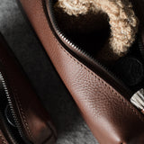 A close-up of a brown pebbled leather zip pouch shown open, with a beige sponge visible inside. A second matching pouch lies partly out of frame on the left, and the main pouch contains small accessories like a toothbrush and a small brush.