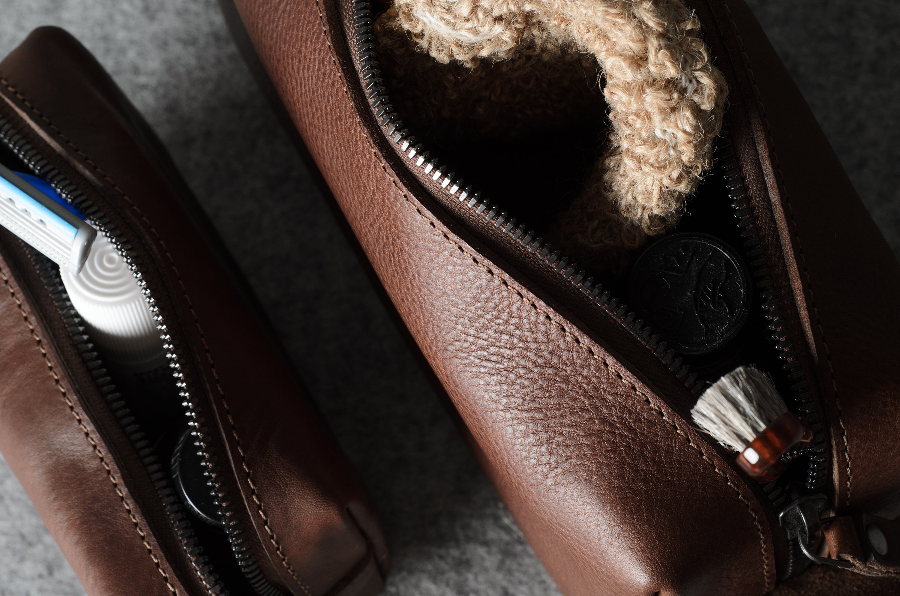 A close-up of a brown pebbled leather zip pouch shown open, with a beige sponge visible inside. A second matching pouch lies partly out of frame on the left, and the main pouch contains small accessories like a toothbrush and a small brush.