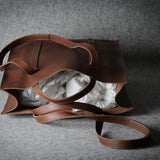 A brown leather tote bag with a matching shoulder strap, shown open with white tissue paper inside, set against a textured gray background.