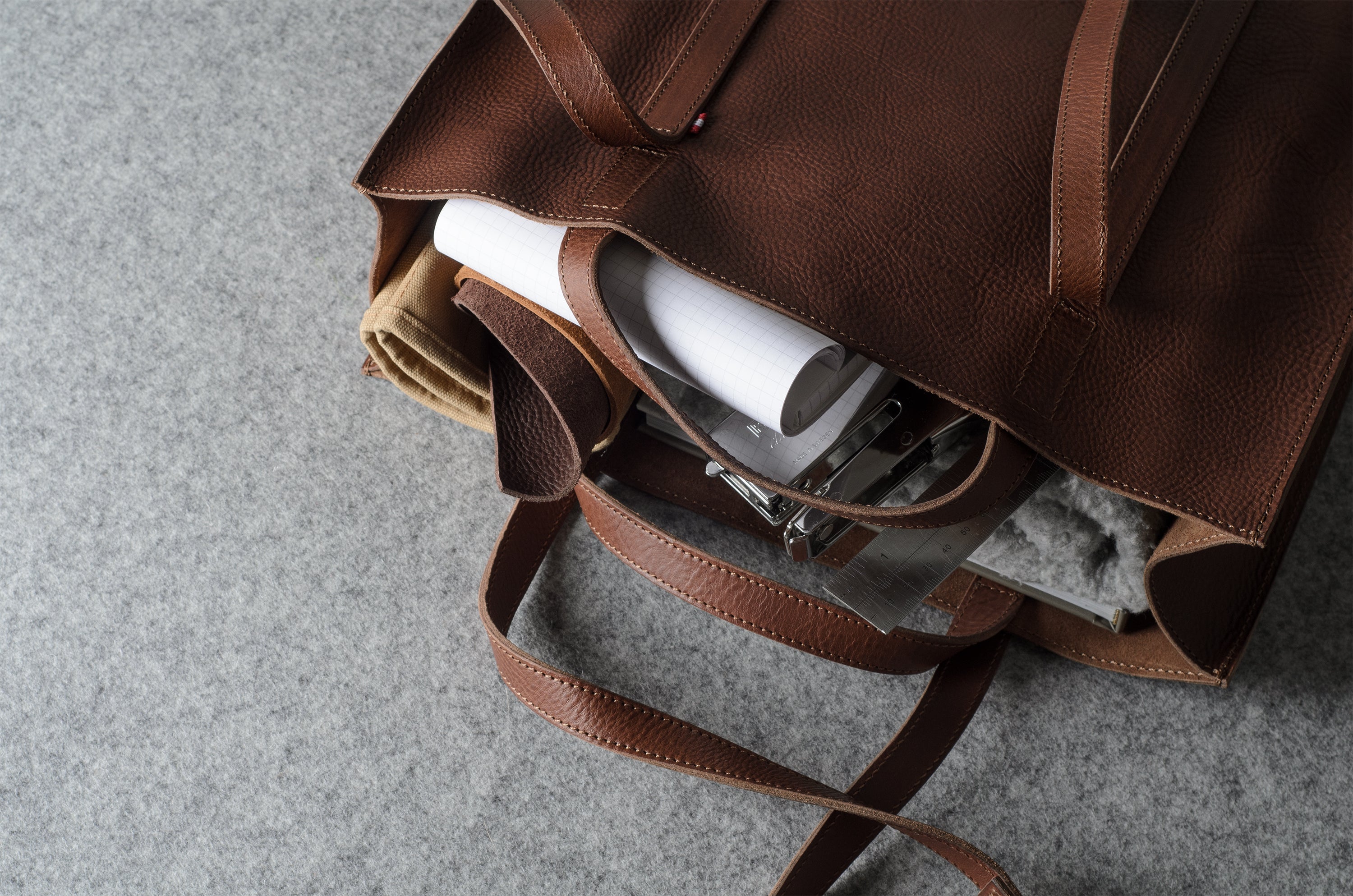 A brown pebbled leather tote bag shown from an overhead angle, partially open to reveal a white cylindrical item, a beige fabric piece, and a gray textured insert inside, set against a gray felt background.