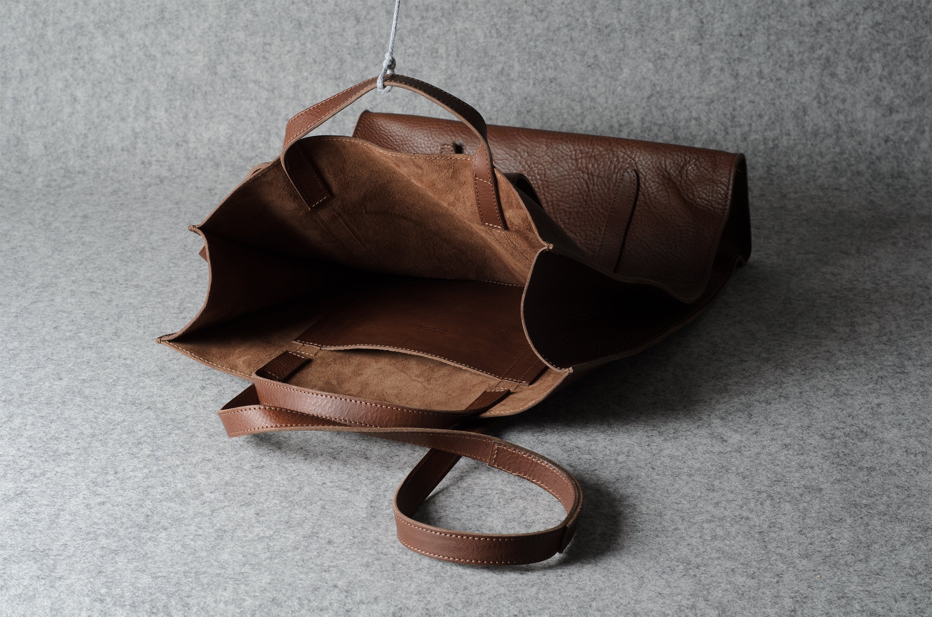 A brown leather tote bag with a matching shoulder strap, shown open against a textured gray background.
