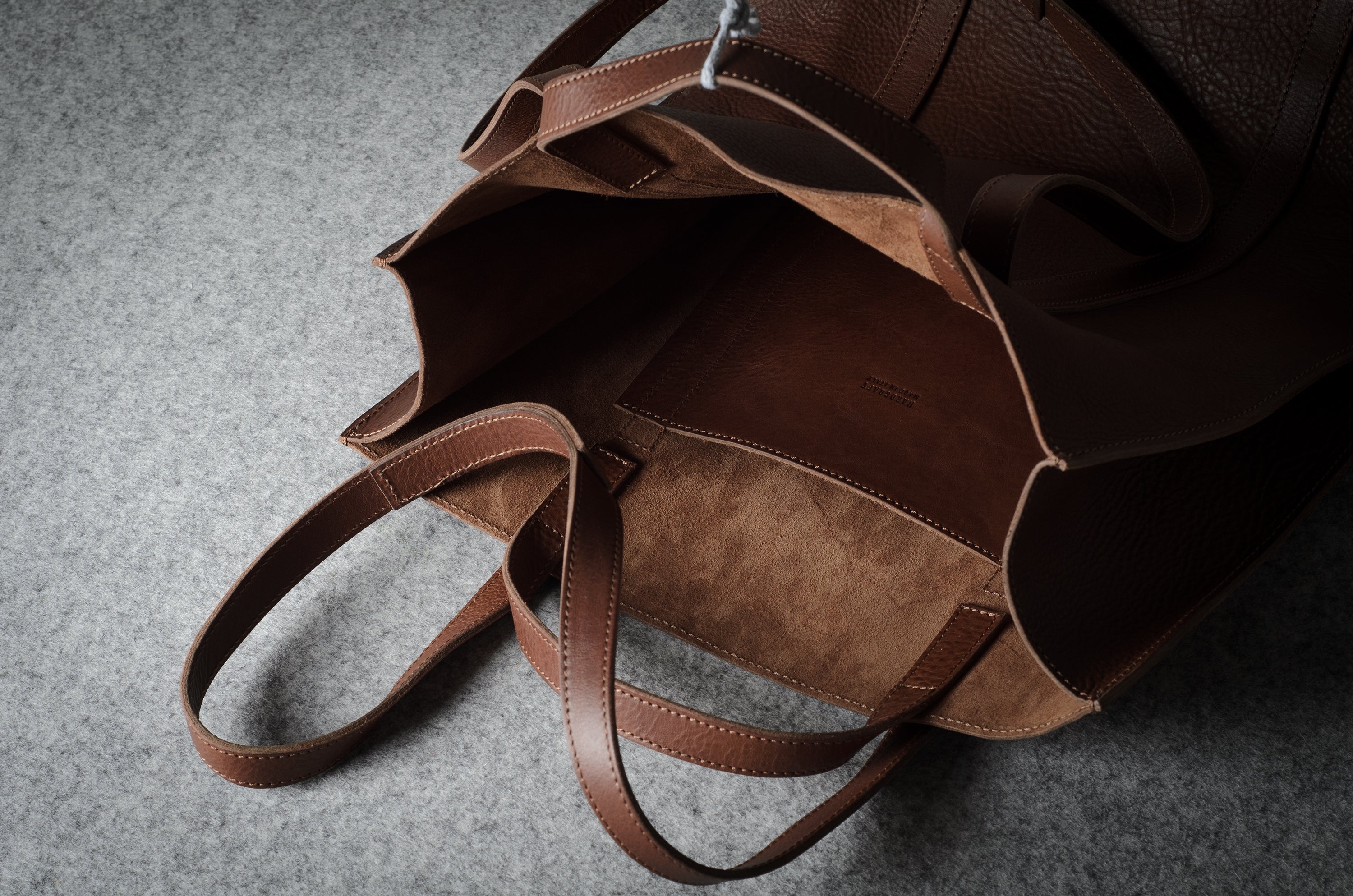A close-up of a brown leather tote bag shown open, highlighting its dual-texture exterior and interior lining on a gray textured surface.