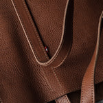 Close-up of a brown pebbled leather tote bag showing the texture, stitching, and a small red brand tab on the strap.