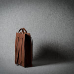 A brown leather tote bag with integrated handles, standing upright on a gray textured studio floor against a matching gray backdrop.
