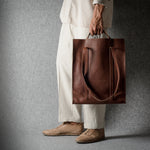 A person wearing a cream-colored outfit and tan shoes holds a large brown leather tote bag against a dark gray textured studio background.