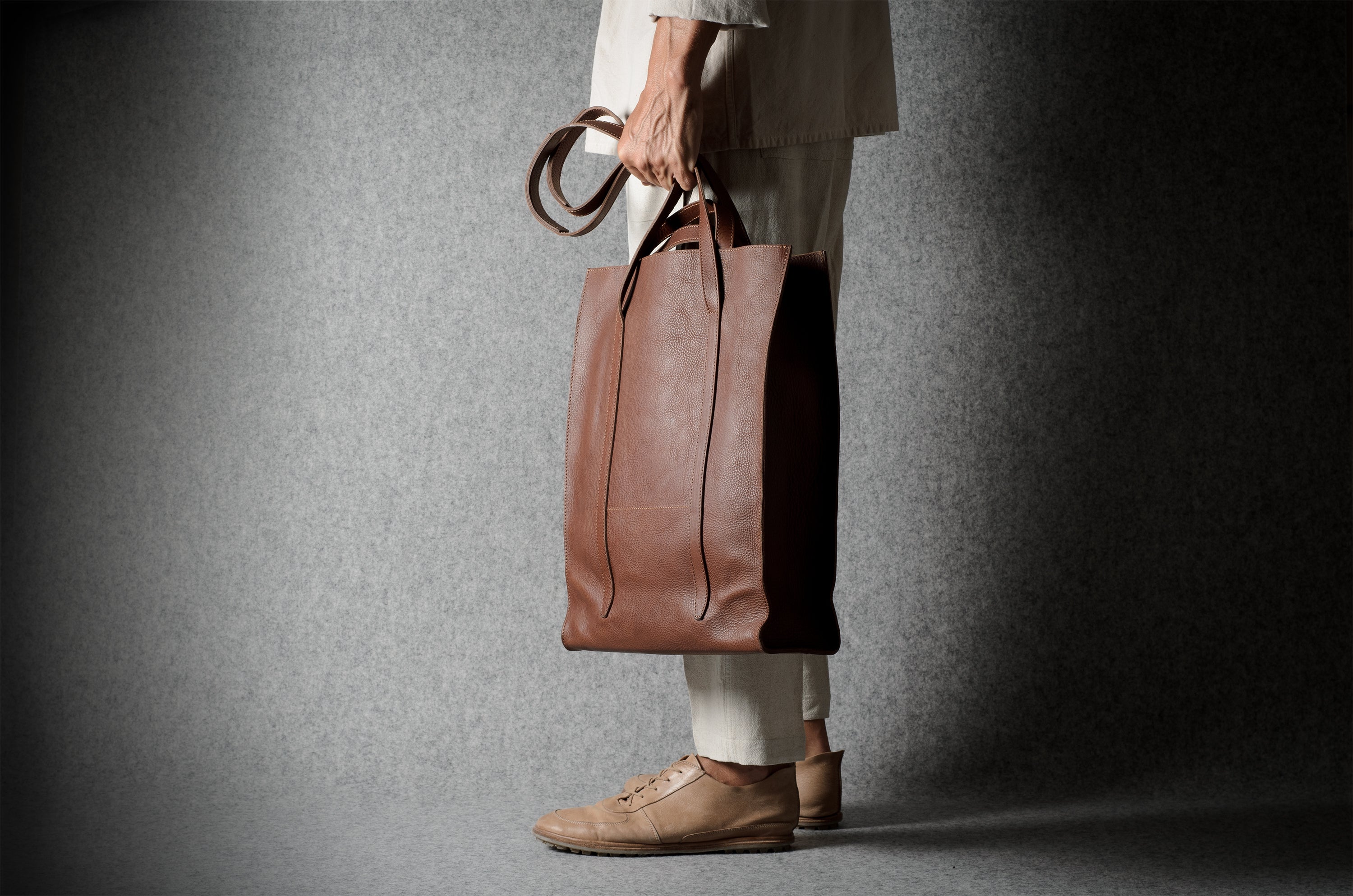 A person in a beige blazer and white trousers holds a large brown leather tote bag against a dark gray textured studio backdrop.