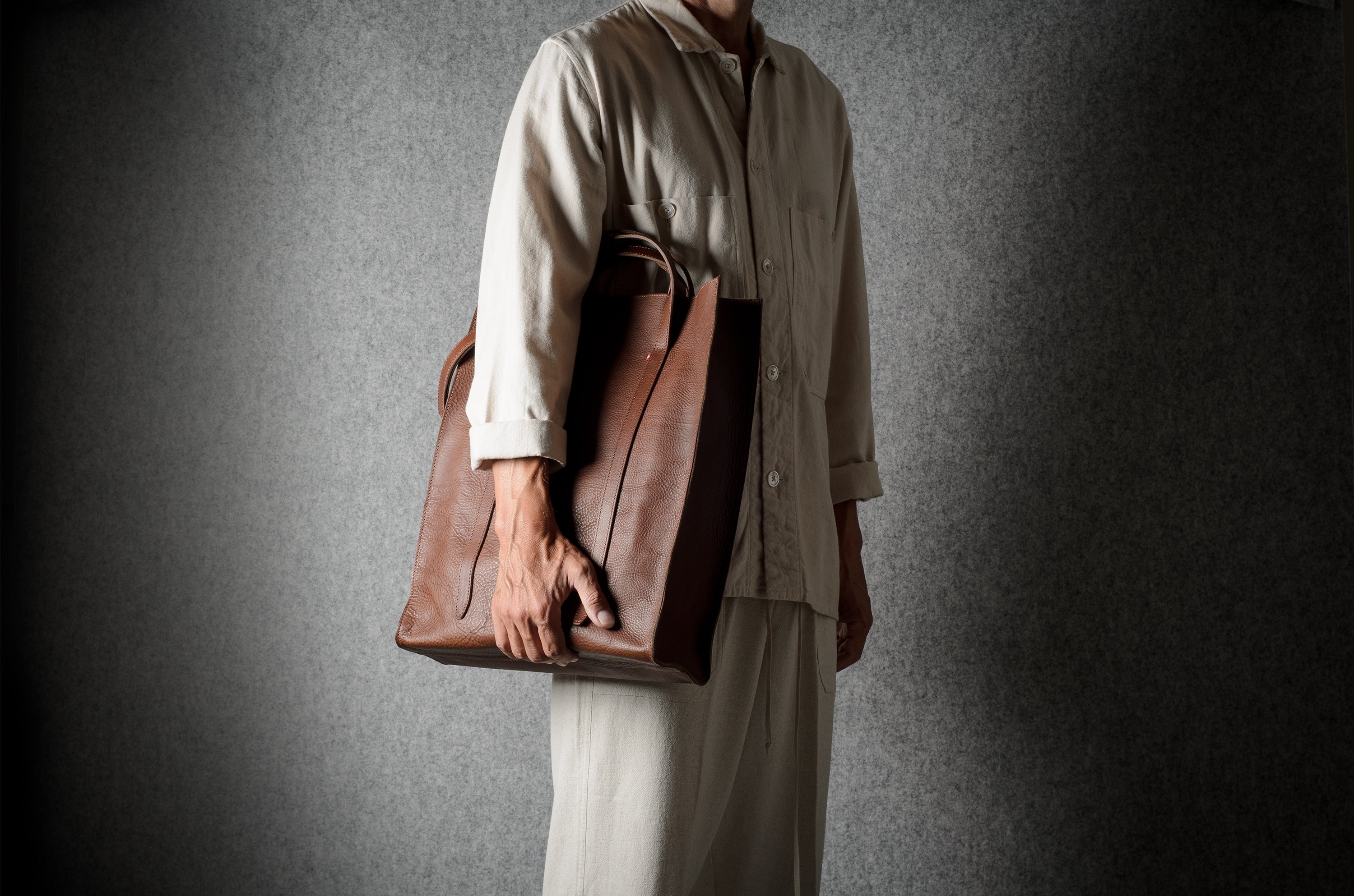 A person wearing a light beige traditional outfit holds a large brown leather tote bag against a dark gray textured background.