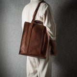 A person wearing a cream-colored outfit is shown from behind, carrying a large brown leather tote bag over their shoulder against a dark textured gray wall.