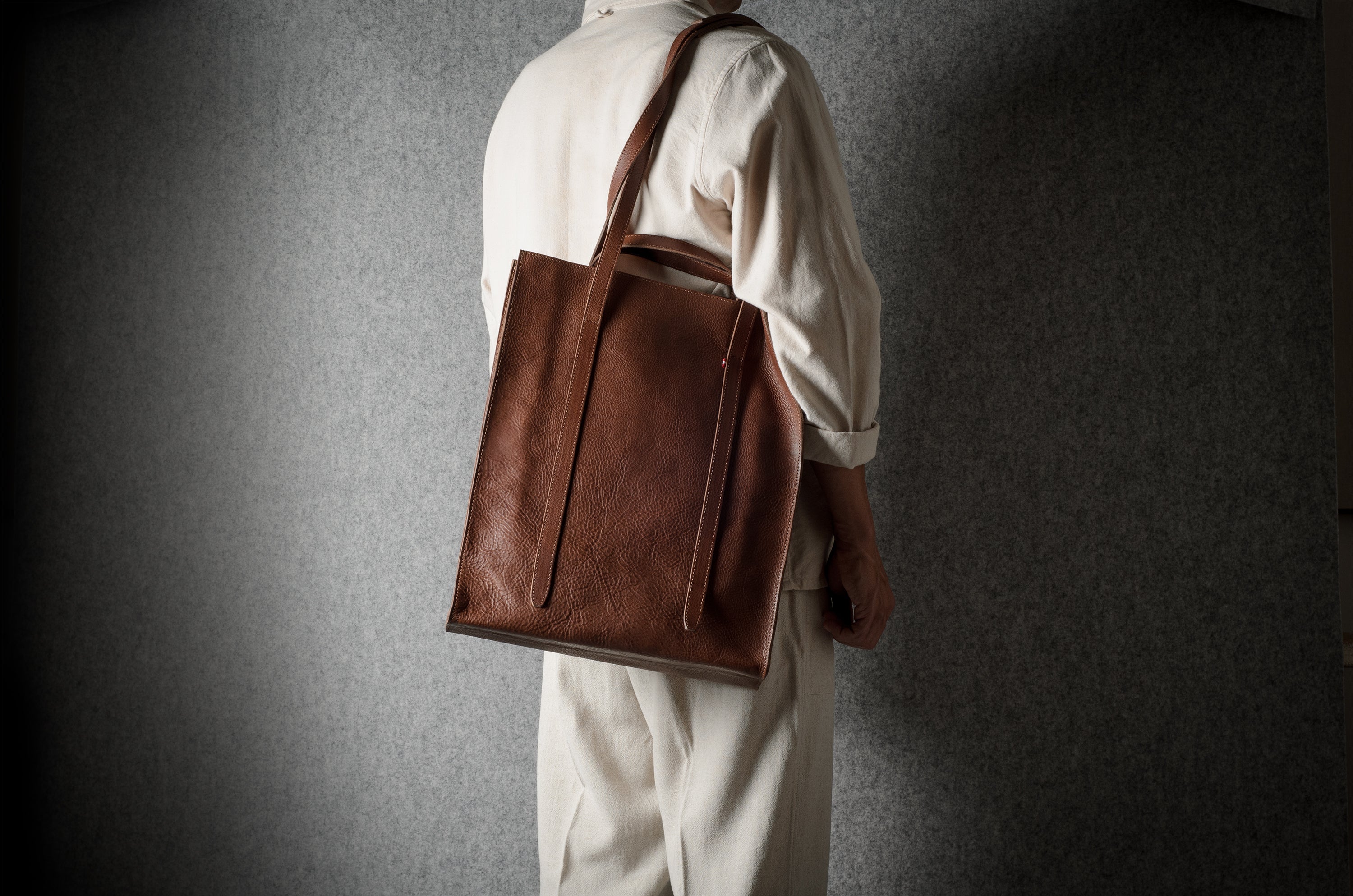 A person wearing a cream-colored outfit is shown from behind, carrying a large brown leather tote bag over their shoulder against a dark textured gray wall.