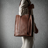 A person in a cream-colored outfit carries a large brown leather tote bag against a textured gray background.