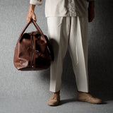 A person wearing a light beige shirt and wide-leg trousers holds a brown leather duffel bag against a textured gray studio backdrop.