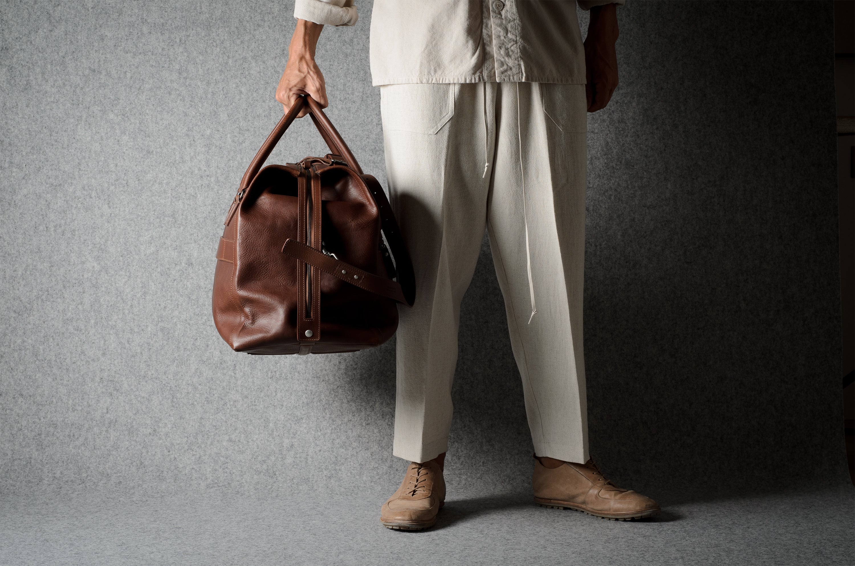 A person wearing a light beige shirt and wide-leg trousers holds a brown leather duffel bag against a textured gray studio backdrop.