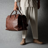 A person wearing light beige clothing holds a brown leather duffel bag against a dark gray textured studio backdrop.