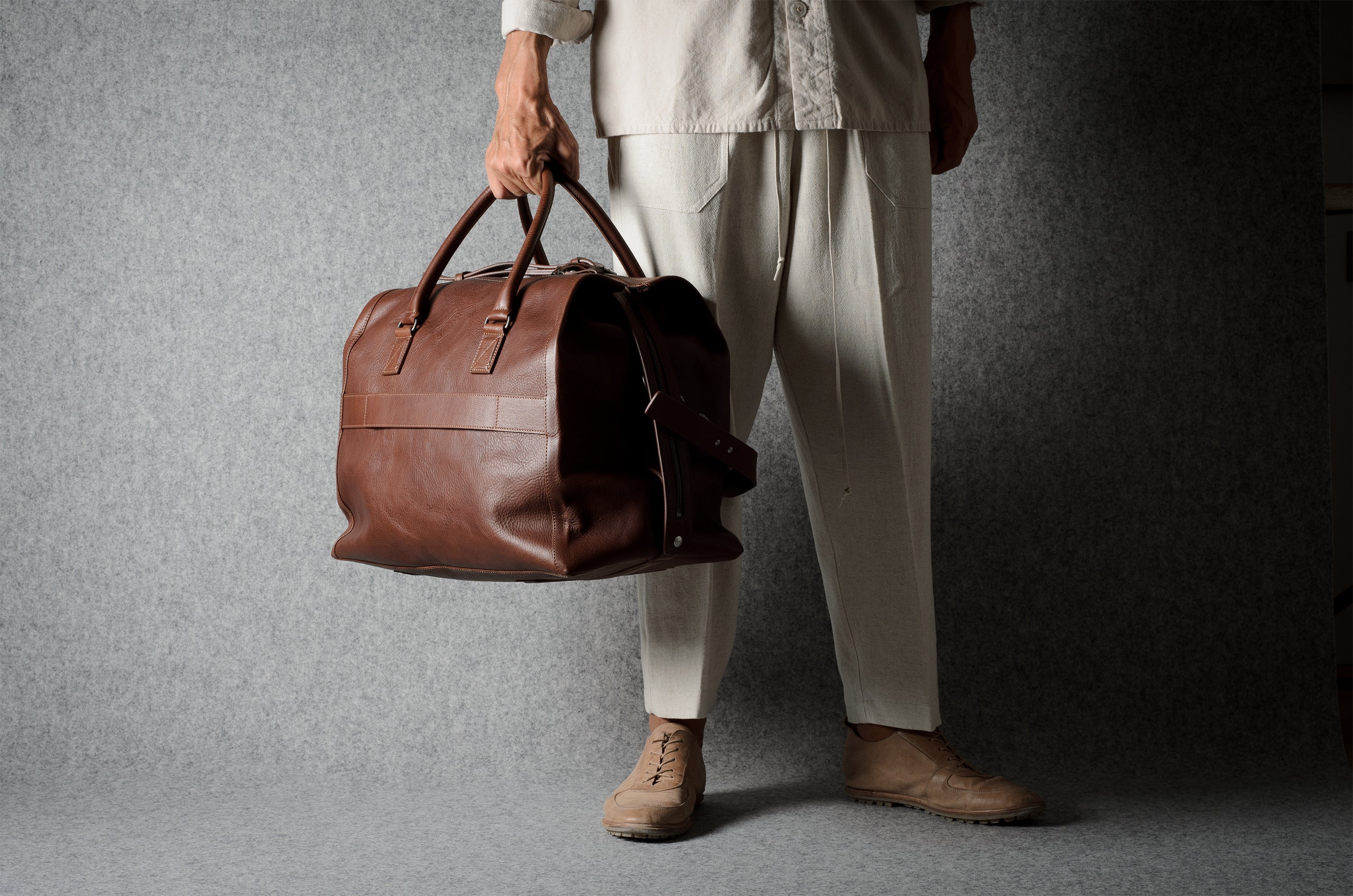A person wearing light beige clothing holds a brown leather duffel bag against a dark gray textured studio backdrop.