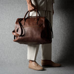A person wearing light beige trousers and tan shoes holds a brown leather duffel bag against a dark gray textured background.