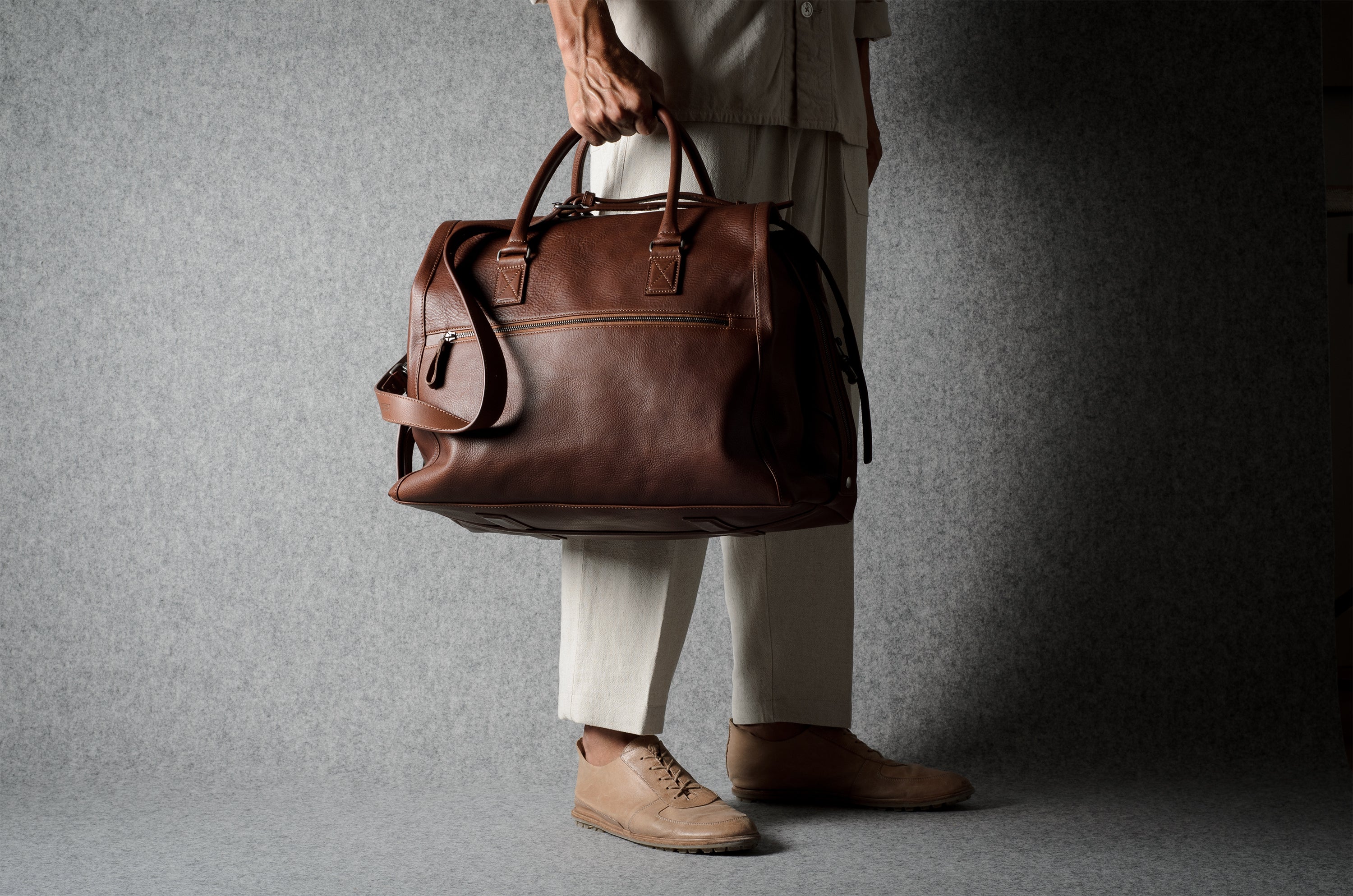 A person wearing light beige trousers and tan shoes holds a brown leather duffel bag against a dark gray textured background.