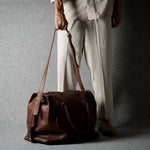 A person wearing light beige trousers and a matching top holds a brown leather duffel bag against a textured gray studio backdrop.