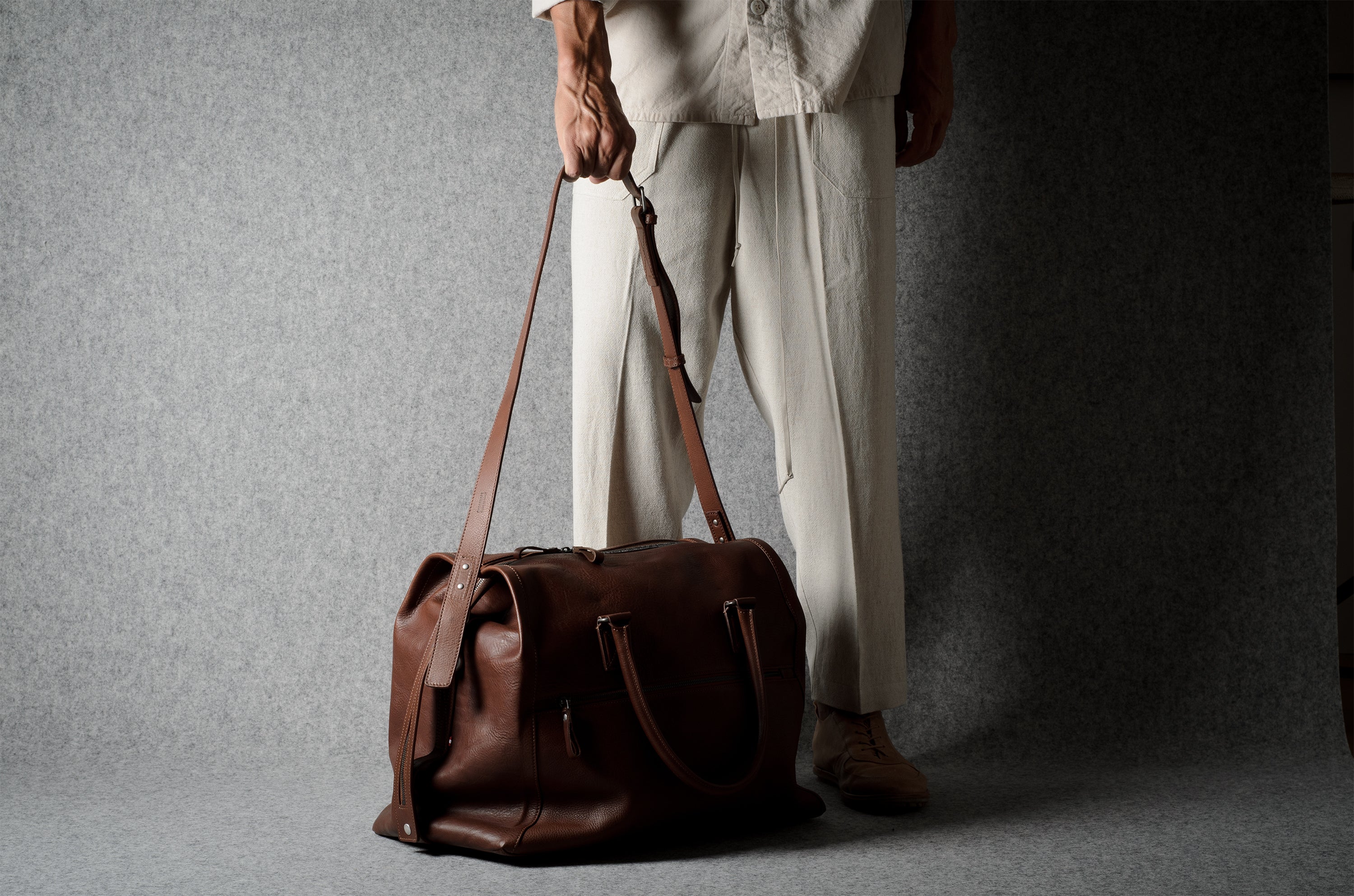 A person wearing light beige trousers and a matching top holds a brown leather duffel bag against a textured gray studio backdrop.