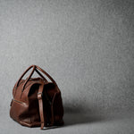 A brown leather duffel bag with dual top handles and a detachable shoulder strap, positioned on a textured gray studio background.