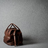 A brown leather duffel bag with dual top handles and a detachable shoulder strap, positioned on a textured gray studio background.
