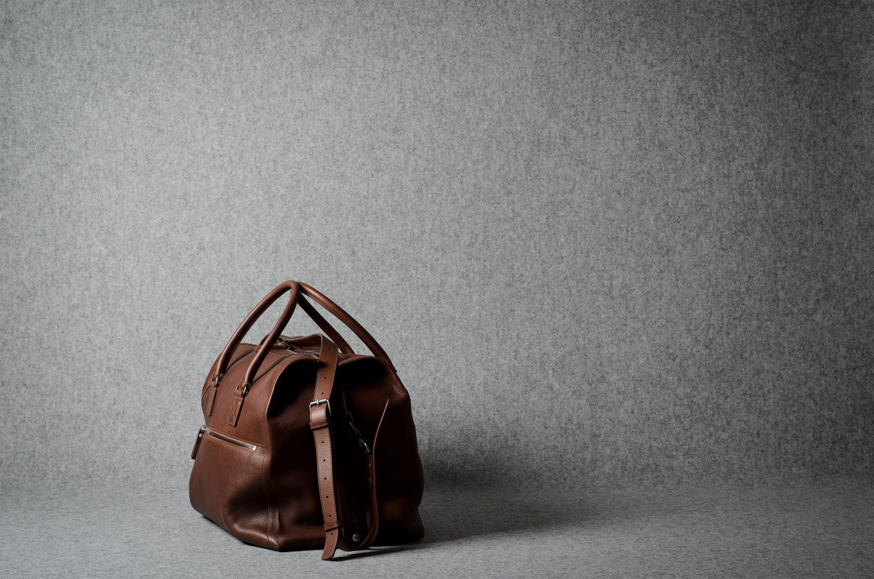 A brown leather duffel bag with dual top handles and a detachable shoulder strap, positioned on a textured gray studio background.