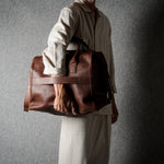 A person in a light beige shirt and trousers carries a large brown leather duffel bag against a dark gray textured background.