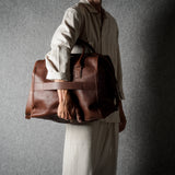 A person in a light beige shirt and trousers carries a large brown leather duffel bag against a dark gray textured background.