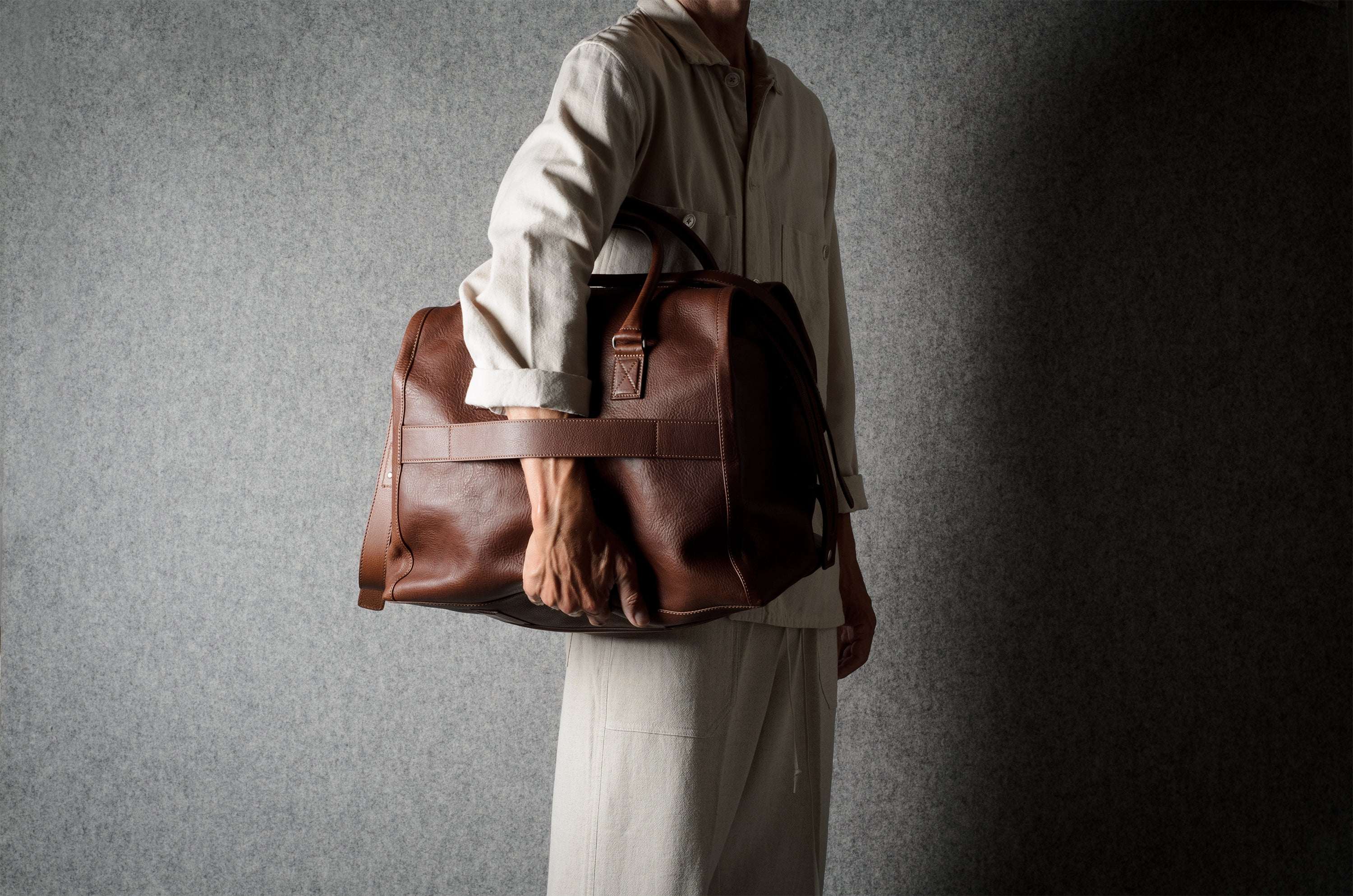A person in a light beige shirt and trousers carries a large brown leather duffel bag against a dark gray textured background.