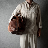 A person wearing a light beige shirt and pants holds a brown leather duffel bag against a dark gray textured background.