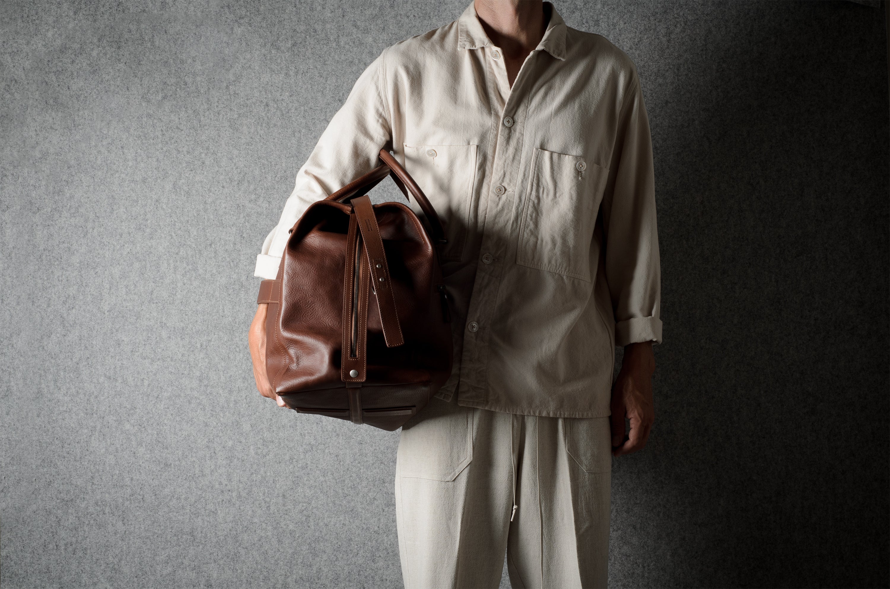 A person wearing a light beige shirt and pants holds a brown leather duffel bag against a dark gray textured background.