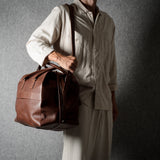 A person wearing a light beige shirt and trousers carries a brown leather duffel bag against a dark textured gray background.