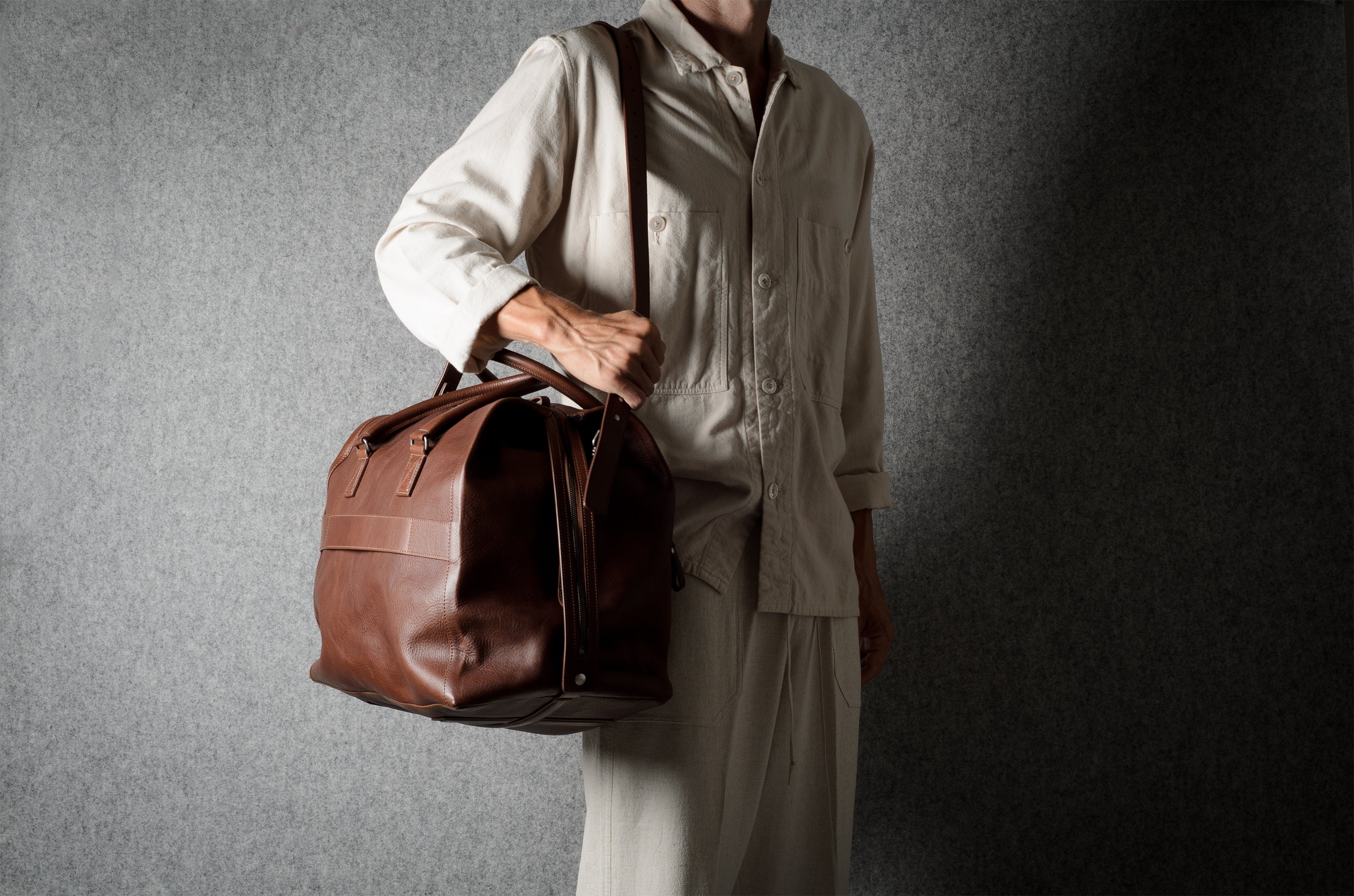 A person wearing a light beige shirt and trousers carries a brown leather duffel bag against a dark textured gray background.